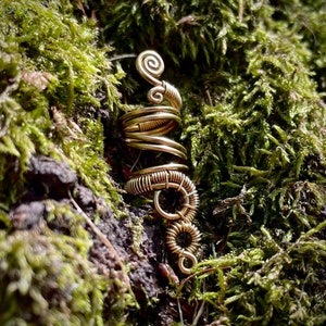 Puede incluir: Un pendiente de alambre dorado con un diseño en espiral. El pendiente está hecho de múltiples bobinas de alambre que están retorcidas juntas. El pendiente está colocado sobre un fondo de musgo verde.