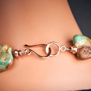 May include: Close-up of a copper clasp on a turquoise gemstone necklace. The clasp is a simple loop with a hook closure. The turquoise stones are a mottled green and brown colour.