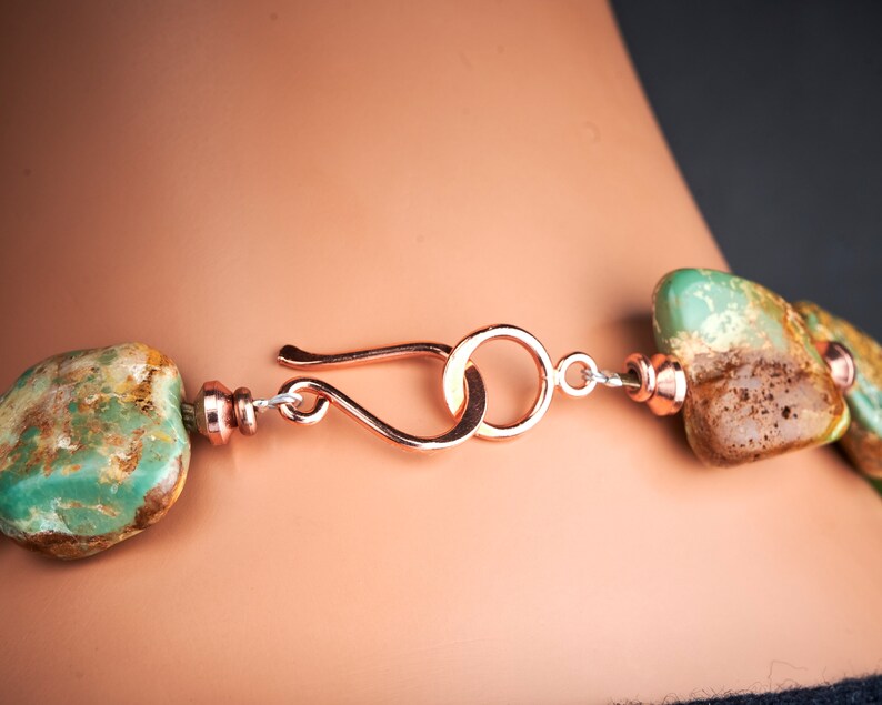 May include: Close-up of a copper clasp on a turquoise gemstone necklace. The clasp is a simple loop with a hook closure. The turquoise stones are a mottled green and brown colour.