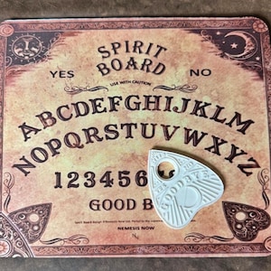 May include: A vintage-style Spirit Board with the words "SPIRIT BOARD" and "YES" and "NO" printed on it. The board features the alphabet, numbers, and a planchette. The planchette is white and has the words "GOODBYE" and "YES" and "NO" printed on it.