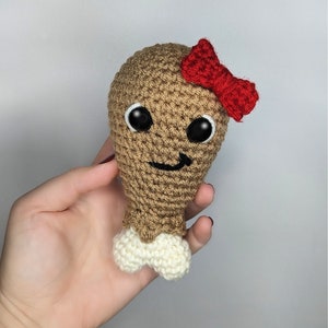 May include: A crocheted chicken leg toy with a red bow and embroidered eyes. The toy is brown and white and has a smiling face.