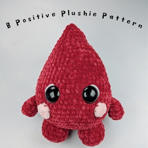 Puede incluir: Un peluche rojo hecho a mano con ojos negros grandes, brazos y pies pequeños. El peluche tiene forma de lágrima y pequeñas mejillas rosadas. El texto encima del peluche dice "B Positive Plushie Pattern".