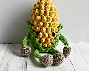 May include: A crocheted corn on the cob with green leaves and brown feet. The corn has a cute face with black eyes.
