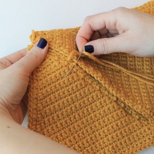 May include: A close-up of a person's hands sewing together two pieces of crocheted fabric. The fabric is a mustard yellow color and the person is using a needle and thread to sew the pieces together.