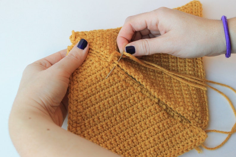 May include: A close-up of a person's hands sewing together two pieces of crocheted fabric. The fabric is a mustard yellow color and the person is using a needle and thread to sew the pieces together.