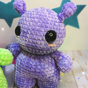 May include: A purple crocheted plush toy with large black eyes and a small pink nose. The toy has a rounded body and two small ears.