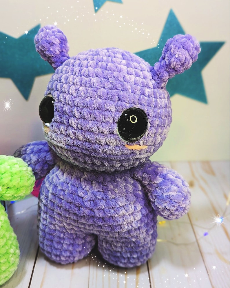 May include: A purple crocheted plush toy with large black eyes and a small pink nose. The toy has a rounded body and two small ears.