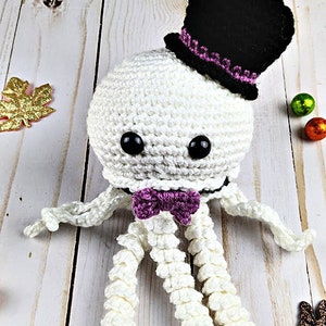 Puede incluir: Pulpo de crochet blanco con un sombrero de copa negro y una pajarita morada. El pulpo tiene ojos negros y está sentado sobre una superficie de madera blanca.