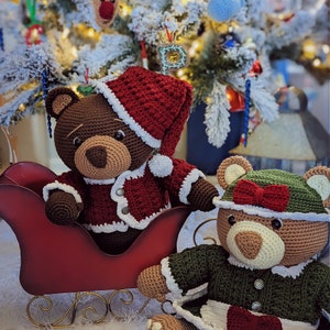 May include: Two crocheted teddy bears wearing festive outfits. One bear is wearing a red Santa hat and a red and white sweater. The other bear is wearing a green and white sweater with a red bow. The bears are sitting in a red sleigh.