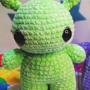 May include: A green crocheted plush toy shaped like an alien with large black eyes. The toy has a textured surface and is standing on a wooden surface.