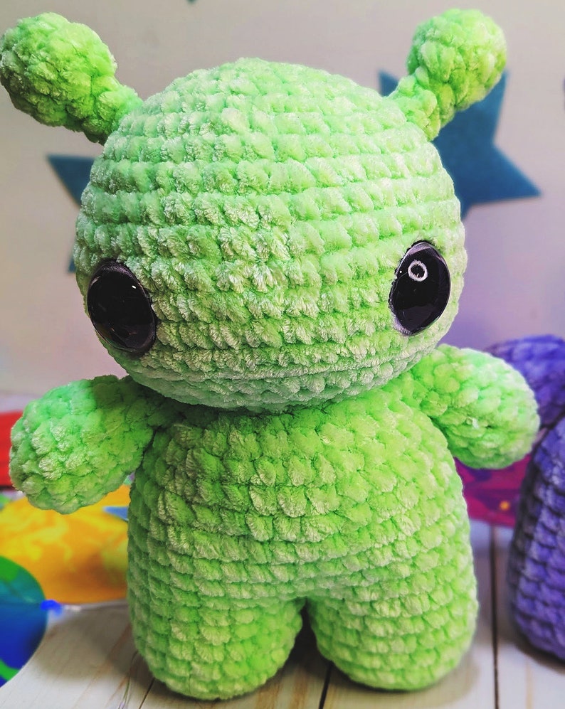 May include: A green crocheted plush toy shaped like an alien with large black eyes. The toy has a textured surface and is standing on a wooden surface.