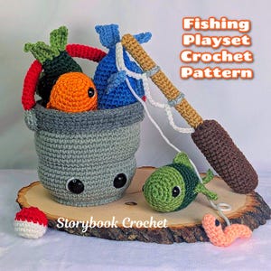 May include: A crochet fishing playset featuring a gray bucket with black eyes, containing a green leafy vegetable, an orange fish, and a blue fish. A brown fishing rod with a white string is attached to a green fish and a pink worm. Text reads Fishing Playset Crochet Pattern and Storybook Crochet.
