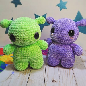 May include: Two crocheted plush toys, one green and one purple, with large eyes and a friendly expression. The toys are made of soft yarn and have a textured surface.