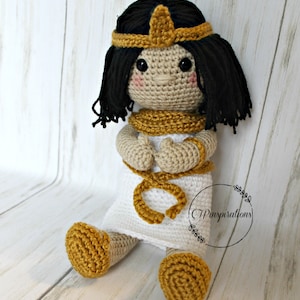 May include: A crocheted doll with black hair, a gold crown, and a white dress. The doll is wearing gold bracelets and a gold necklace. The doll is sitting on a white wooden surface.