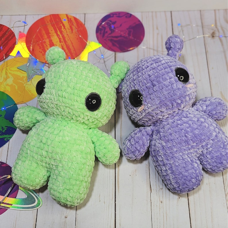 May include: Two crocheted plush toys shaped like aliens. One is green and the other is purple. Both have large eyes and a friendly expression.