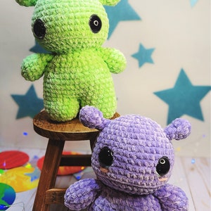 May include: Two crocheted plush toys, one green and one purple, with large black eyes. The toys are sitting on a wooden stool.