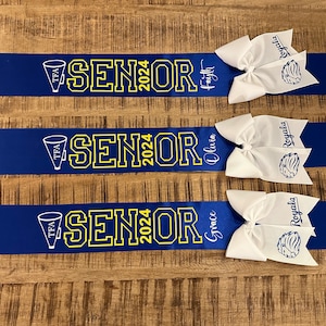 May include: Three blue and yellow graduation sashes with white bows and the text "TFA Senior 2024" and the names "Faith", "Olivia", and "Grace".