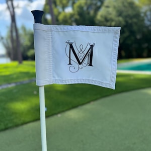May include: A white golf flag with a black decorative letter "M" in the center. The flag is attached to a white pole with a black top. The flag is on a green golf course.