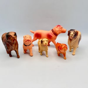May include: A collection of five small dog figurines in various poses and colors. The set includes a dark brown dog, a light tan dog with a black collar, a medium tan dog, a large pinkish-orange dog with a collar, and a tan and brown dog.