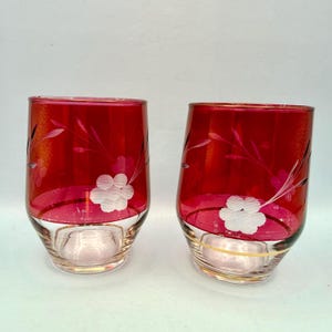 Könnte beinhalten: Zwei rot getönte Trinkgläser mit floralem Design. Die Gläser haben einen klaren Boden mit Goldrand und ein weißes Blumenmuster. Der obere Teil der Gläser ist tiefrot, mit geätzten Blatt- und Stielmotiven.