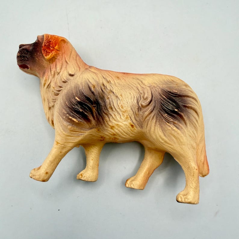 May include: A molded plastic dog figurine featuring a cream-colored body with dark brown patches on the face, shoulders, and hindquarters. The figure depicts a long-haired canine standing in profile with a textured coat and a dark brown muzzle.