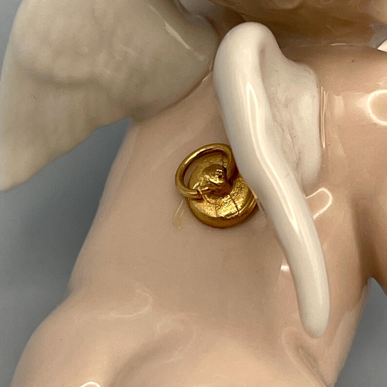 May include: A close-up of a porcelain figurine with a gold-colored metal attachment. The figurine has a pale pink body, white wings, and a gold-colored metal ring. The wings are smooth and curved, and the metal attachment is textured.