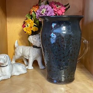 May include: A dark blue ceramic vase with a speckled glaze. The vase has a rounded body and a flared lip. It is sitting on a wooden shelf with two white porcelain figurines.
