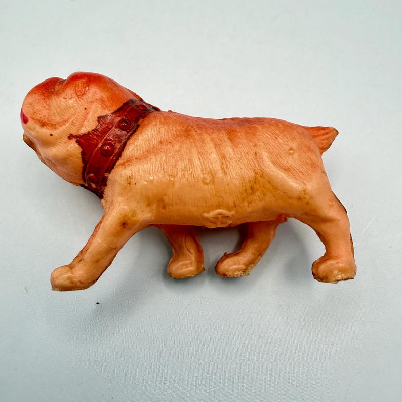 May include: A small, molded orange plastic figurine of a bulldog in a standing position. The dog features a textured surface, a short tail, and a dark red collar with circular stud details around its neck. The facial features are molded.
