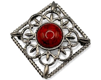 Op de afbeelding: Een vintage broche met een ruitvormig ontwerp. De broche heeft een centrale, ronde, rode steen, omgeven door een sierlijk, zilverkleurig metalen frame. Het frame heeft een gedetailleerd, opengewerkt patroon met een gedraaide touwrand.