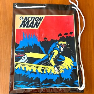 Könnte beinhalten: Ein schwarzer Action Man-Kordelzugbeutel mit einer farbenfrohen Illustration eines Tauchers in einem Boot. Der Taucher trägt einen gelb-blauen Tauchanzug. Die Tasche hat einen weißen Kordelzug und das Action Man-Logo.