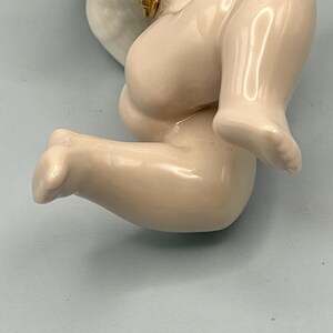 May include: A close-up of a porcelain cherub figurine, featuring a kneeling pose. The cherub has a smooth, off-white finish with subtle shading. The details include a rounded leg and foot, with a hint of a golden accent on the wing.