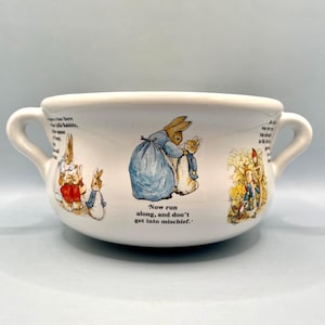 Puede incluir: Cuenco de cerámica blanco con dos asas, con ilustraciones de personajes de Peter Rabbit. El cuenco tiene forma redondeada e incluye el texto: «Ahora corre, y no te metas en líos».