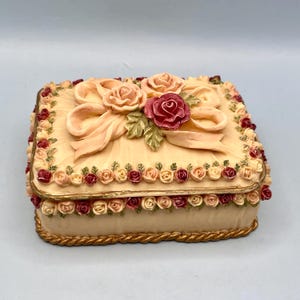 May include: A square, cream-colored trinket box with a decorative floral design. The lid features a bow and roses in shades of pink and red. The box is trimmed with small roses and a gold rope-like border.