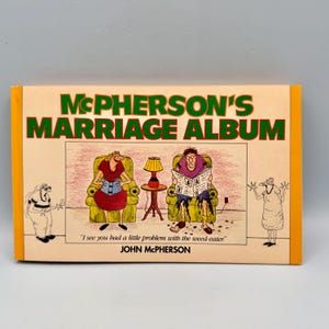 Puede incluir: Un libro titulado "McPherson's Marriage Album" con una ilustración de dibujos animados. La portada presenta el título en letras verdes, una escena de dibujos animados de una pareja y el nombre del autor. El libro tiene una columna vertebral naranja.