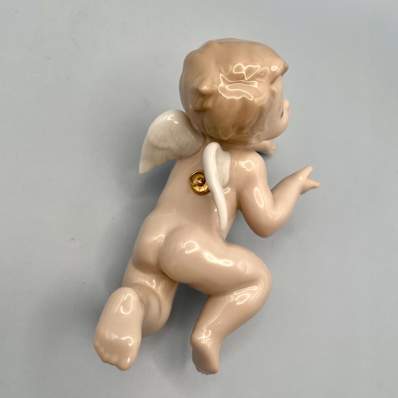 May include: A porcelain figurine of a cherub in a kneeling pose, with small white wings and a golden accent on its back. The cherub has a light skin tone and is set against a light blue background.