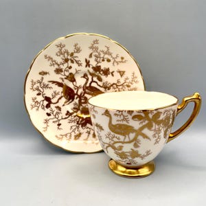 May include: A white teacup and saucer set with gold accents. The cup and saucer feature a gold bird and floral design. The rim and handle of the cup are also gold. The saucer has a scalloped edge.
