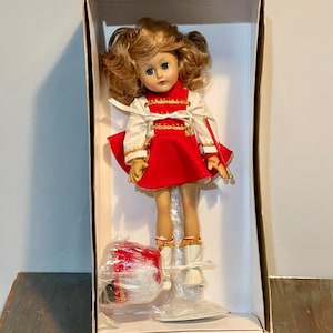 Puede incluir: Una muñeca de porcelana vestida con un uniforme de banda de marcha rojo y blanco con ribetes dorados. La muñeca sostiene una batuta roja y tiene un tambor delante de ella.