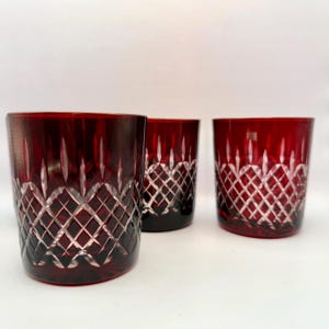 May include: Three ruby red crystal glasses with a diamond pattern. The glasses have a thick base and a straight, slightly tapered shape. The cut crystal design reflects light, creating a sparkling effect.