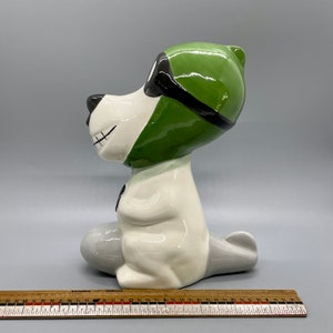 Puede incluir: Una figurita de cer&aacute;mica blanca de un perro con un gorro de aviador verde y gafas. El perro est&aacute; sentado con las piernas cruzadas y tiene una cola gris.