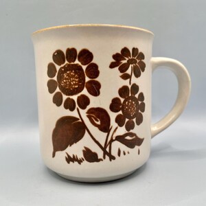 May include: Cream-colored ceramic mug with a brown floral design. The flowers are stylized with detailed petals and leaves. The mug has a rounded handle and a gold rim.
