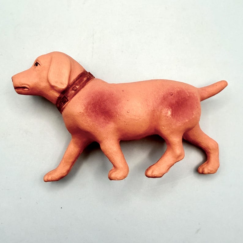 May include: A small, solid-colored peach-toned dog figurine in a standing position. The figure features a textured surface, a molded collar around the neck, and darker reddish-brown patches on the shoulders and hindquarters, facing to the left.