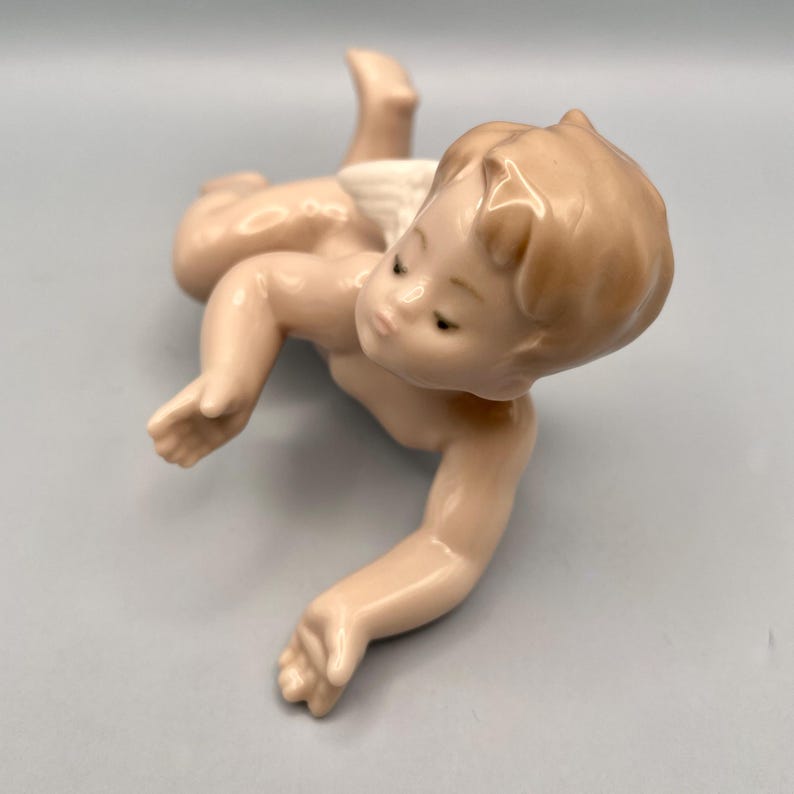 May include: A porcelain figurine of a baby crawling, with a light skin tone and a small white diaper. The baby has a serene expression and is posed on a light gray surface. The figurine has a glossy finish.
