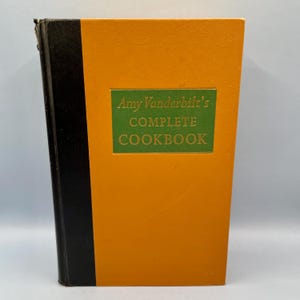 May include: A vintage cookbook titled "Amy Vanderbilt's COMPLETE COOKBOOK." The book has a bright orange cover with a green rectangular label. The title is in gold lettering. The spine is black.