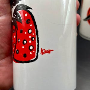 K&ouml;nnte beinhalten: Wei&szlig;e Keramiktasse mit einer schwarzen und roten Illustration einer Erdbeere. Die Tasse ist signiert mit "LeMieux".