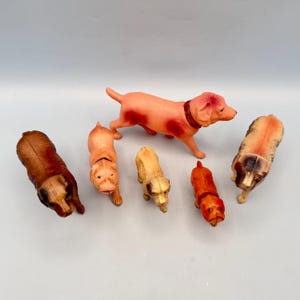 May include: A collection of five small molded plastic dog figurines in various poses and colors. The set includes a large pinkish-tan dog, a dark brown dog, a light tan dog, a smaller cream-colored dog, and a small reddish-brown dog.