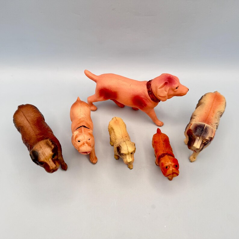 May include: A collection of five small molded plastic dog figurines in various poses and colors. The set includes a large pinkish-tan dog, a dark brown dog, a light tan dog, a smaller cream-colored dog, and a small reddish-brown dog.