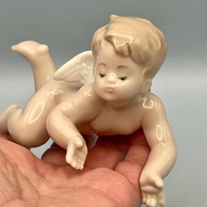 May include: A porcelain cherub figurine in a crawling pose, with a serene expression. The cherub has a light skin tone, with delicate wings and detailed facial features. The figurine is held in a hand, showcasing its size and craftsmanship.