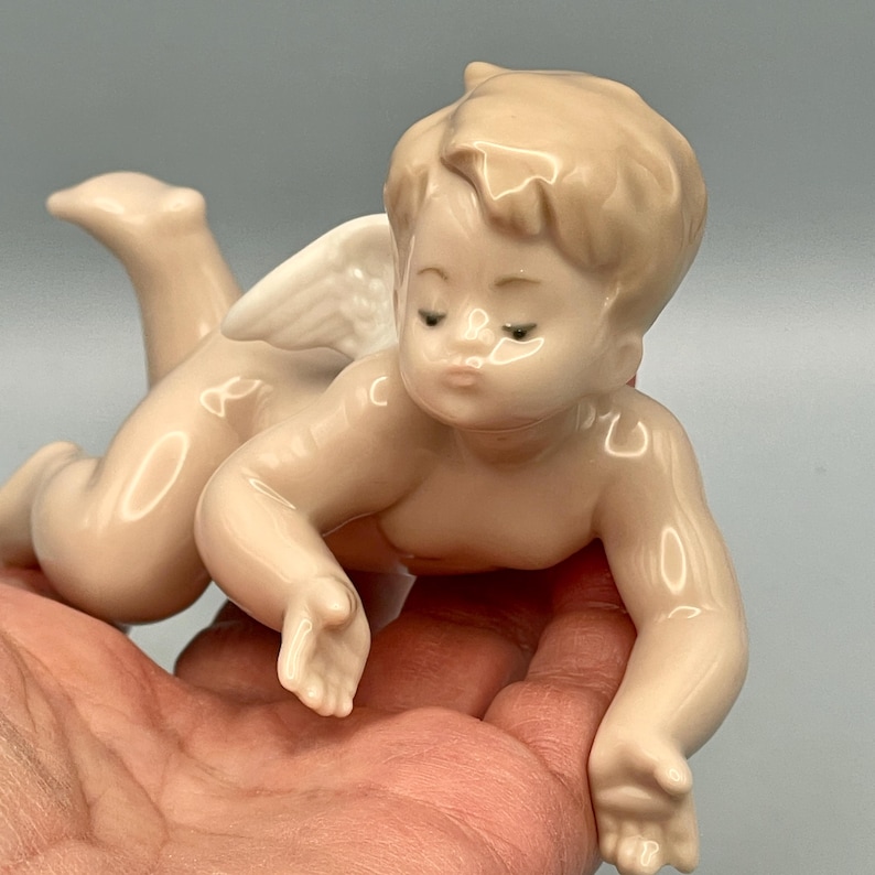 May include: A porcelain cherub figurine in a crawling pose, with a serene expression. The cherub has a light skin tone, with delicate wings and detailed facial features. The figurine is held in a hand, showcasing its size and craftsmanship.