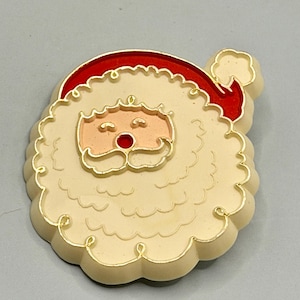 May include: A Santa Claus enamel pin featuring a cream-coloured beard and moustache, a peach-toned face with closed eyes, a bright red nose, and a red hat with a cream-coloured pom-pom. The design is outlined in gold metal with a raised, textured surface.