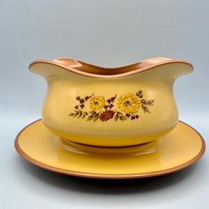 May include: A yellow ceramic gravy boat with a matching saucer, featuring a brown rim and a central floral pattern of yellow and brown flowers with green leaves. The gravy boat has a wide, curved pouring spout and a smooth, glossy finish.
